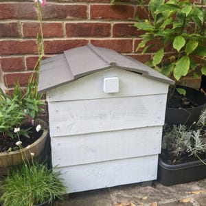 May include: A light grey wooden structure with a dark grey gabled roof and a small white square block centred on the front panel. The structure is constructed from horizontal wooden planks joined with visible screws on the sides.