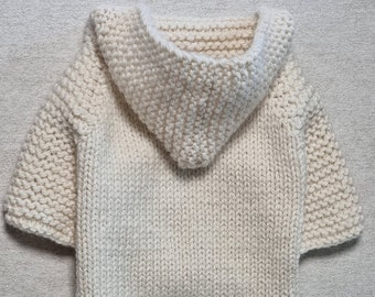 May include: Cream-colored, hand-knitted dog sweater with a hood. The sweater features a textured knit pattern and short sleeves. The hood is a prominent feature, adding a cozy element to the garment. The overall design is simple and functional.