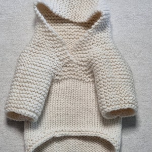 May include: Cream-colored, hand-knitted dog sweater with a hood. The sweater features a V-neck design and sleeves. The knitwear is made with a thick yarn, creating a textured appearance. The dog sweater is designed to keep a small dog warm.