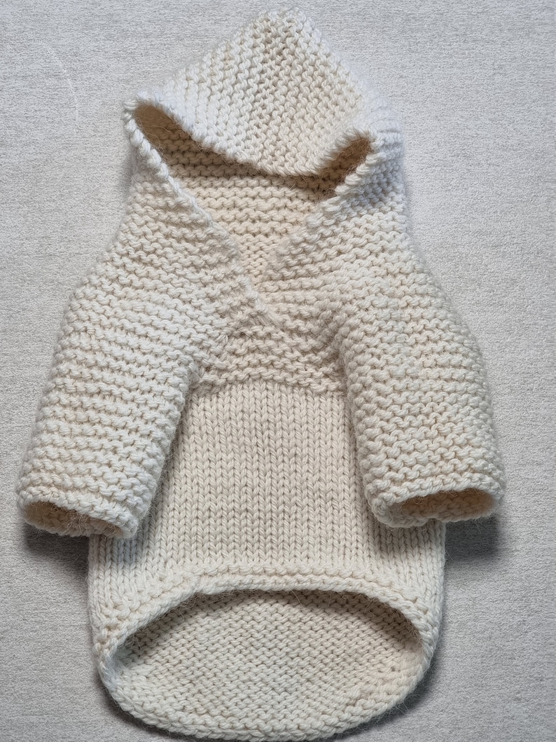 May include: Cream-colored, hand-knitted dog sweater with a hood. The sweater features a V-neck design and sleeves. The knitwear is made with a thick yarn, creating a textured appearance. The dog sweater is designed to keep a small dog warm.