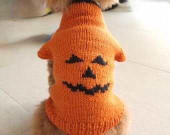 May include: An orange knitted dog jumper featuring a black jack-o'-lantern face pattern on the back. The garment has a ribbed collar and hem, with short sleeves and a textured knit construction, worn by a small, light brown, curly-haired dog.