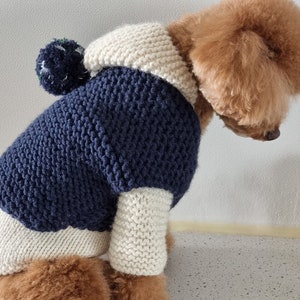 May include: A navy blue and cream-colored knitted dog sweater with a pom-pom on the hood. The sweater has a textured knit pattern and is designed to fit a small dog. The dog is a light brown color.
