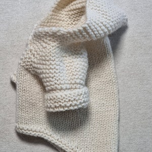 May include: Cream-colored knitted baby sweater with a hood and one sleeve visible. The sweater is made of thick yarn and has a textured appearance. The hood is folded over, and the sleeve is rolled up.