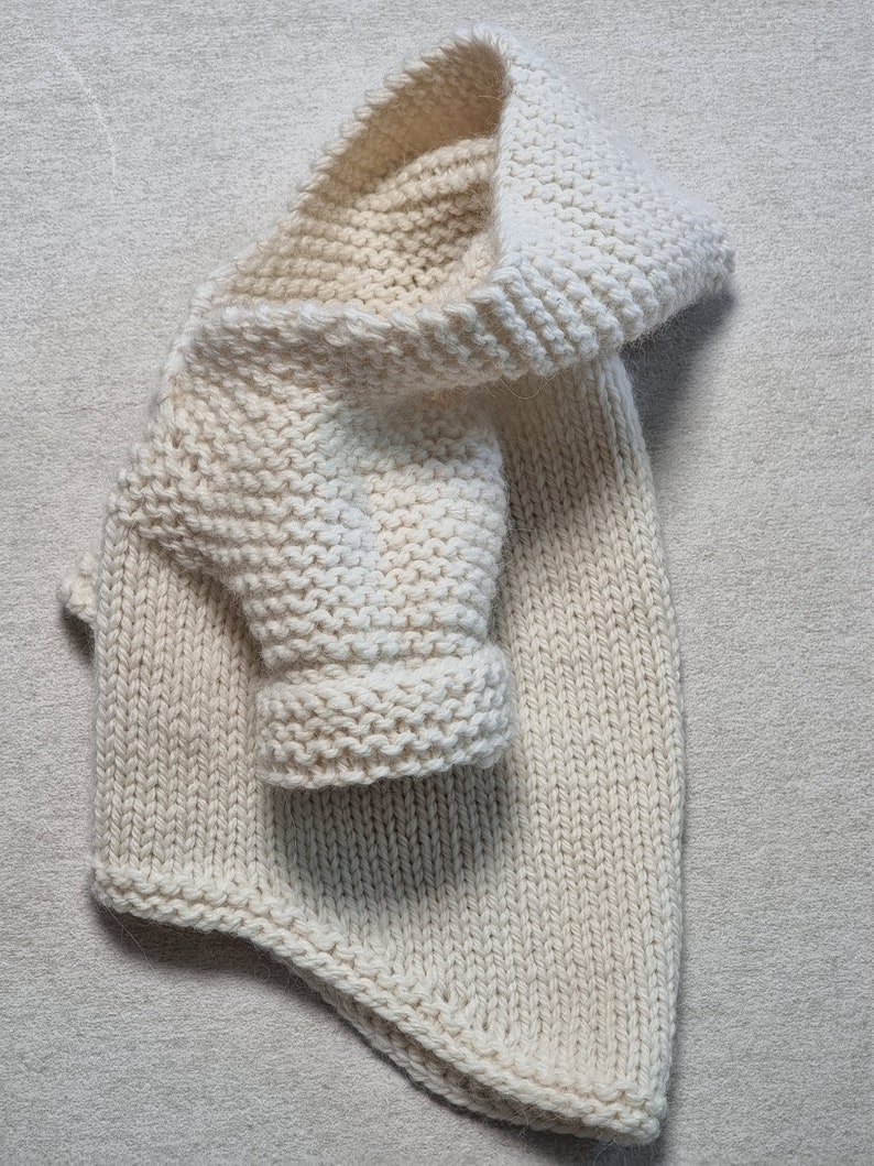 May include: Cream-colored knitted baby sweater with a hood and one sleeve visible. The sweater is made of thick yarn and has a textured appearance. The hood is folded over, and the sleeve is rolled up.