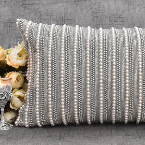 May include: A silver and white beaded throw pillow with a decorative design. The pillow is made of a soft fabric and has a luxurious feel.