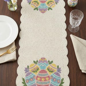 May include: Beige beaded Easter table runner with scalloped edges. The runner features colorful beaded Easter egg designs in pink, yellow, blue, and purple. A vase of flowers, a plate, and a glass are also on the table.