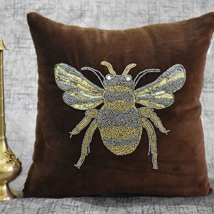 May include: A brown velvet pillow with a beaded bee design. The bee is made of gold and silver beads and has a detailed design.