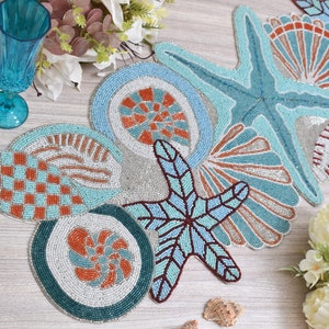 Może przedstawiać: Bielony bieżnik stołowy z koralików z motywem muszli i rozgwiazd. Bieżnik wykonany jest z białych, niebieskich i pomarańczowych koralików i przedstawia różne muszle i rozgwiazdy.