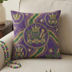 May include: Square purple beaded pillow with gold, green, and white accents. The pillow features a central design of a crown, mask, and musical instruments, surrounded by circles with similar motifs. Additional crowns and masks are scattered across the surface. A strand of beads is visible.