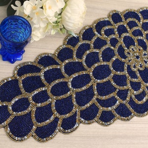 Op de afbeelding: Een blauwe en gouden kralen tafelrunner met een bloemmotief. De runner is gemaakt van kleine kralen die aan elkaar zijn genaaid om een ingewikkeld patroon te creëren.