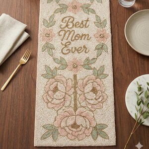 May include: Beaded table runner with the text "Best Mom Ever" surrounded by pink and green floral designs. The runner is beige with a floral pattern in pink, green, and gold. A gold fork and beige napkin are on the left.
