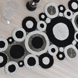 Peut inclure: Un chemin de table en perles noires et blanches avec un motif circulaire. Le chemin de table est fait de petites perles cousues ensemble pour créer un design unique.