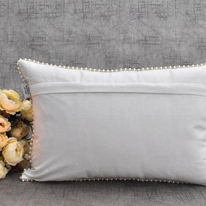 Puede incluir: Almohada blanca con un ribete de perlas alrededor de los bordes. La almohada est&aacute; tendida sobre una superficie gris texturizada.