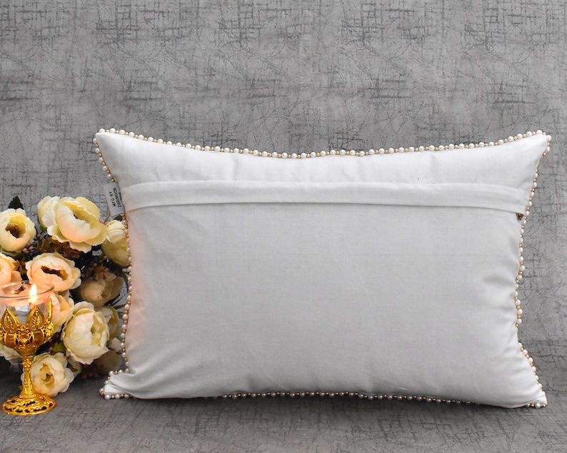 Puede incluir: Almohada blanca con un ribete de perlas alrededor de los bordes. La almohada est&aacute; tendida sobre una superficie gris texturizada.