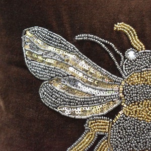 May include: A brown velvet pillow with a large, embroidered bee design. The bee is made of silver and gold sequins and beads.