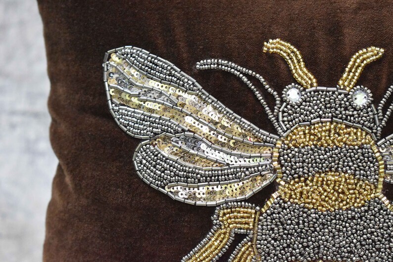 May include: A brown velvet pillow with a large, embroidered bee design. The bee is made of silver and gold sequins and beads.