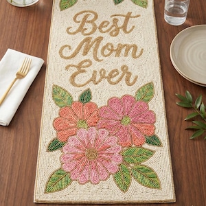 Puede incluir: Camino de mesa con cuentas con el texto dorado "Best Mom Ever". Presenta tres grandes flores en tonos rosa y coral, con hojas verdes. El fondo es crema y los bordes están ribeteados en dorado.