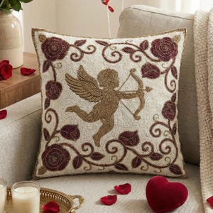 May include: Beige square pillow with a beaded design featuring a gold Cupid with a bow and arrow, surrounded by burgundy roses and vines. The pillow has a gold beaded border and is on a beige sofa.