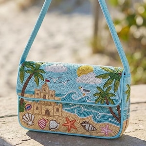 May include: A beaded handbag with a beach scene design. The bag features a light blue background with a sandcastle, palm trees, seagulls, and seashells. The strap and edges are light blue. The bag is a rectangle shape.