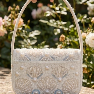 May include: A white handbag featuring intricate beadwork in the shape of seashells. The bag is adorned with small white seed beads, larger faux pearls, and subtle light blue accents. It has a single top handle and a structured rectangular shape.