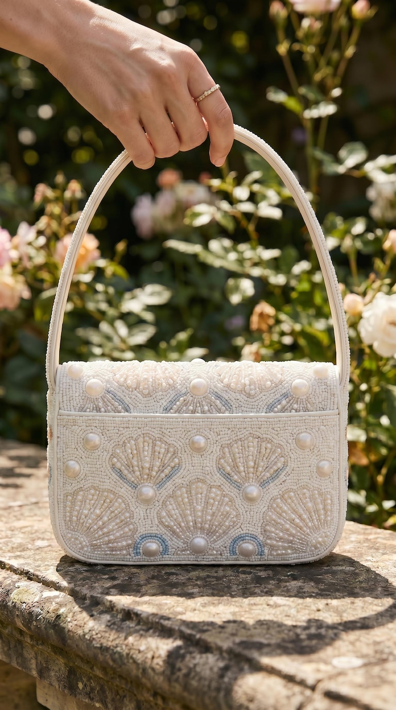May include: A white handbag featuring intricate beadwork in the shape of seashells. The bag is adorned with small white seed beads, larger faux pearls, and subtle light blue accents. It has a single top handle and a structured rectangular shape.