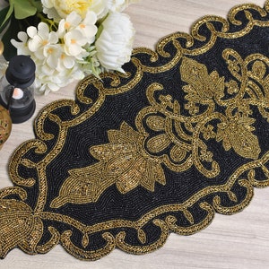 Op de afbeelding: Zwarte kralen tafelloper met gouden bloemen- en krulwerkontwerp. De loper heeft een geschulpte rand en is versierd met gouden kralen. De loper is ongeveer 30 cm breed en 183 cm lang.