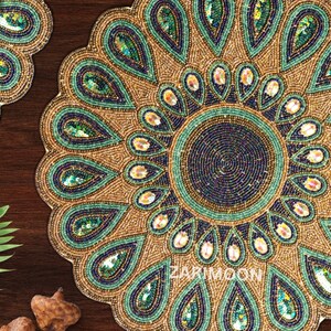May include: A round beaded table mat with a peacock design. The mat is made of gold, green, and blue beads. The center of the mat has a circular design with a smaller circle in the center. The mat is 12 inches in diameter.