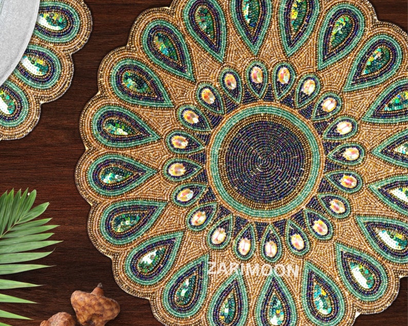 May include: A round beaded table mat with a peacock design. The mat is made of gold, green, and blue beads. The center of the mat has a circular design with a smaller circle in the center. The mat is 12 inches in diameter.