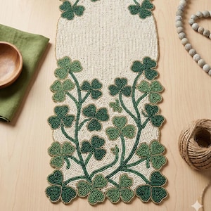 以下が含まれることがあります： 緑のクローバーのデザインが施された、ビーズで作られたアイボリーのテーブルランナー。 クローバーは緑の茎に沿って配置され、装飾的なボーダーを形成しています。 木製のボウルと緑のリネンナプキンが背景に見えます。