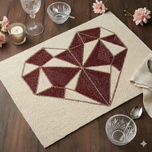 May include: Beige placemat with a geometric heart design. The heart is made of burgundy and cream-colored beads. The placemat is on a wooden table, with a candle, glassware, and a small bowl.