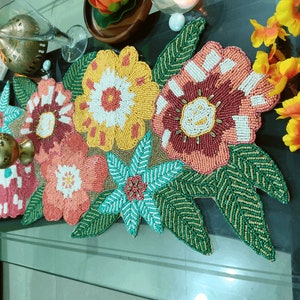 Puede incluir: Un camino de mesa de cuentas con un diseño floral. El camino de mesa presenta flores rosas, amarillas, verdes y azules con acentos blancos. Las flores están rodeadas de hojas verdes.