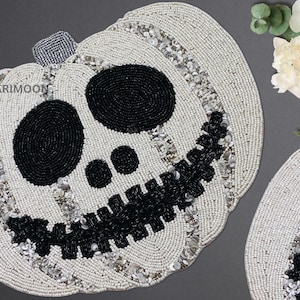 May include: White beaded pumpkin placemat with a black skull face design. The pumpkin has a stitched mouth and bead accents.
