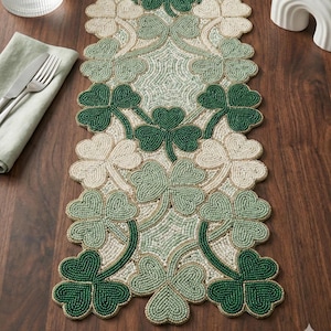 May include: A beaded table runner with a repeating shamrock design in various shades of green and cream. The shamrocks are outlined with gold beads, offering a decorative touch. Ideal for adding a festive touch to a dining table.