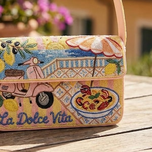 K&ouml;nnte beinhalten: Eine Perlenhandtasche mit einem rosa Riemen und einem Klappverschluss. Die Tasche hat ein farbenfrohes Design mit einem Roller, Zitronen, einem Sonnenschirm und einer Sch&uuml;ssel Pasta. Die Worte "La Dolce Vita" sind auf der Vorderseite zu sehen.
