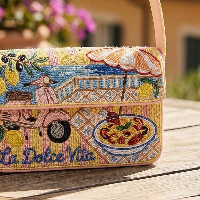 K&ouml;nnte beinhalten: Eine Perlenhandtasche mit einem rosa Riemen und einem Klappverschluss. Die Tasche hat ein farbenfrohes Design mit einem Roller, Zitronen, einem Sonnenschirm und einer Sch&uuml;ssel Pasta. Die Worte "La Dolce Vita" sind auf der Vorderseite zu sehen.