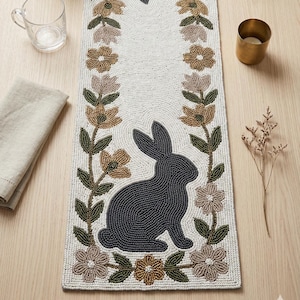 Può includere: Runner da tavola pasquale con perline con una silhouette di coniglio grigio scuro e accenti floreali marroni, verdi e rosa. Il runner è bianco con una trama di perline, ideale per l'arredamento primaverile.