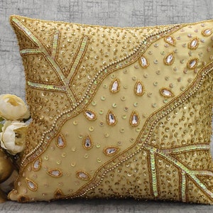 May include: A gold throw pillow with a beaded design. The pillow has a light gold background with a pattern of sequins and beads in a teardrop shape. The pillow is decorated with a gold trim.