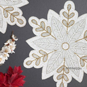 May include: White beaded snowflake-shaped placemat with gold leaf accents. The placemat is made of white beads and has a gold leaf design. The placemat is perfect for adding a touch of elegance to your table setting.