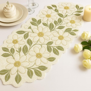 May include: Cream-coloured beaded table runner with a floral design. The runner features large white flowers with yellow centres and green leaves. The edges are scalloped.