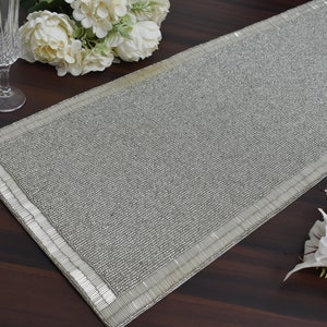 May include: A silver beaded table runner with a white border. The runner is on a wooden table.