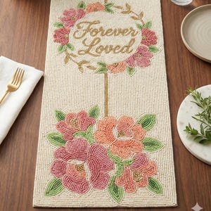 May include: Cream-coloured beaded table runner with a floral design. The runner features a wreath with the words "Forever Loved" and a bouquet of pink and peach flowers. A gold fork and white napkin are on the left.