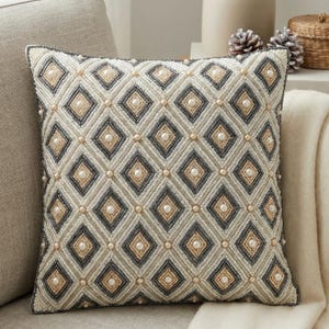 Puede incluir: Cojín decorativo cuadrado con un patrón geométrico de diamantes. Los diamantes son en tonos crema, gris y dorado, con cuentas tipo perla en el centro de cada diamante. El cojín está sobre un sofá de color neutro.