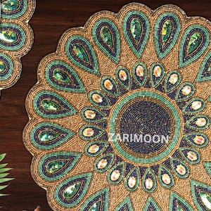 May include: A round beaded placemat with a peacock feather design. The placemat is gold with green and blue beads.