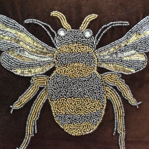 May include: A brown velvet pillow with a large, beaded bee design. The bee is made of silver and gold beads and has a black and yellow striped body.