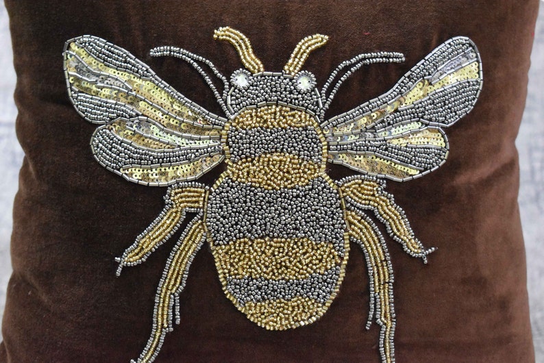 May include: A brown velvet pillow with a large, beaded bee design. The bee is made of silver and gold beads and has a black and yellow striped body.