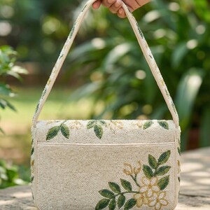May include: Cream-colored beaded handbag with a rectangular shape and a top handle. The bag is decorated with green and gold floral embroidery. The handle and edges are also adorned with beads. The bag is held by a hand against a blurred green background.