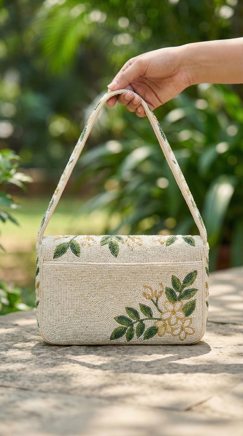 May include: Cream-colored beaded handbag with a rectangular shape and a top handle. The bag is decorated with green and gold floral embroidery. The handle and edges are also adorned with beads. The bag is held by a hand against a blurred green background.