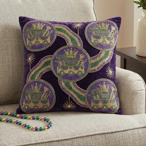 May include: Purple velvet throw pillow with beaded Mardi Gras designs. The pillow features circular beaded designs with crowns, masks, and musical instruments in gold, green, and purple. The pillow is on a neutral-colored sofa.