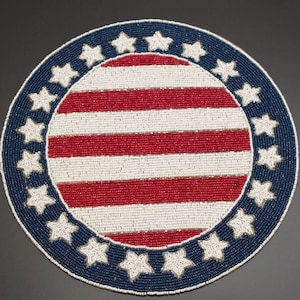 May include: Round beaded placemat with a patriotic design. The center features red and white stripes, surrounded by a blue border with white stars. The beaded design adds texture and detail to the decorative piece.