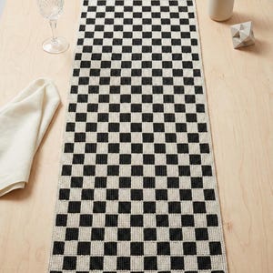 May include: A black and white chequered table runner. The runner is rectangular and features a repeating pattern of black squares on a white background. The runner is displayed on a light wood surface, with a glass, napkin, and decorative items.